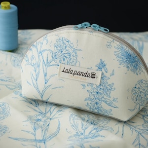 Pode incluir: Uma bolsa de cosméticos branca com um padrão floral azul e um zíper azul claro. A bolsa tem o texto "Lalapanda" em uma pequena etiqueta. Uma bobina de linha azul é visível no fundo.