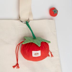 May include: A cream tote bag with a red tomato-shaped pouch attached. The pouch has a green leaf top and a small tag. A smaller tomato-shaped item is also visible. The pouch is attached to the bag with a green string and a metal ring.