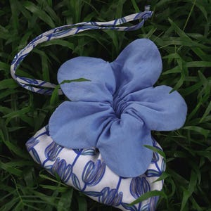 Pode incluir: Uma scrunchie floral azul e uma bolsa de tecido a condizer. A scrunchie tem a forma de uma flor grande. A bolsa é branca com um padrão floral azul e um cordão azul. Os artigos estão sobre relva verde.