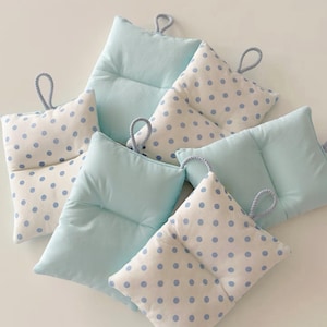 May include: A collection of small, square pillows in light blue and white. Some pillows feature a polka dot pattern in light blue, while others are solid light blue. Each pillow has a loop at the top for hanging.