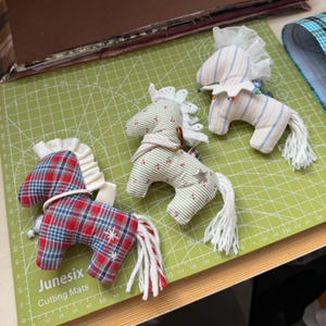 May include: Three handmade fabric horse toys. One is red, blue, and white plaid. Another is green and white striped with a star. The third is white and blue striped. Each has a white mane and tail. Measurements are in centimetres.