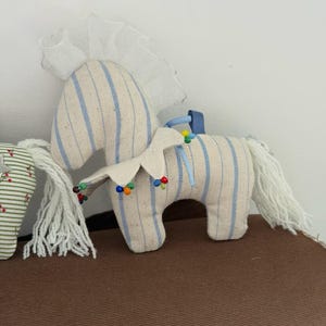 May include: A stuffed toy horse with a cream-colored body and blue vertical stripes. The horse has a white mane and tail, and a decorative collar with colorful beads. A white frilly fabric is attached to the top of the horse's head.