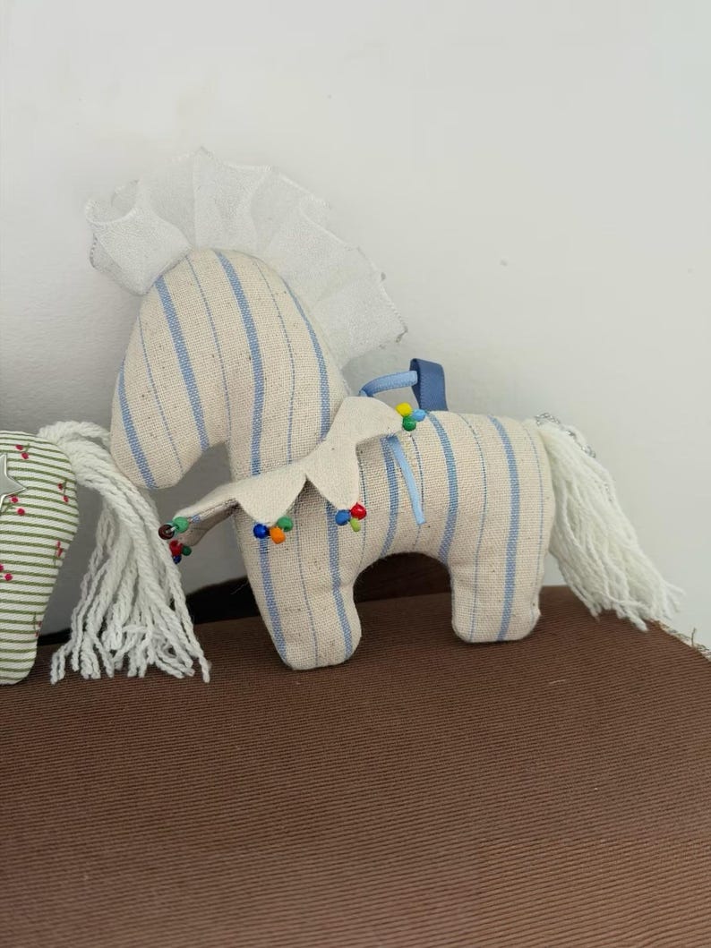 May include: A stuffed toy horse with a cream-colored body and blue vertical stripes. The horse has a white mane and tail, and a decorative collar with colorful beads. A white frilly fabric is attached to the top of the horse's head.