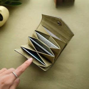 Può includere: Un portafoglio beige in stile origami con più fessure per carte. Il portafoglio è aperto, rivelando diverse carte. L'esterno ha un aspetto strutturato e si vede una chiusura a scatto argentata. Il portafoglio è su una superficie dai toni neutri.