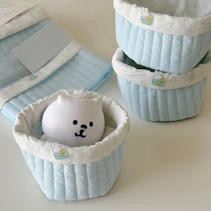 Könnte beinhalten: Ein Set aus hellblauen, gesteppten Stoffkörben mit weißer Einfassung und einem kleinen Hasenaufnäher. Ein Korb enthält einen weißen Plüschbären. Die Körbe sind gestapelt und mit einem passenden gesteppten Stoffstück angeordnet.