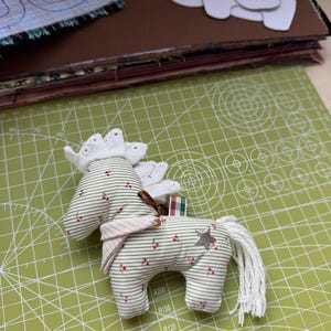 May include: A handmade fabric horse toy with a white mane and tail, adorned with a silver star and red berry accents. The horse is made of striped fabric with a white lace crown and a pink scarf. It rests on a green cutting mat.