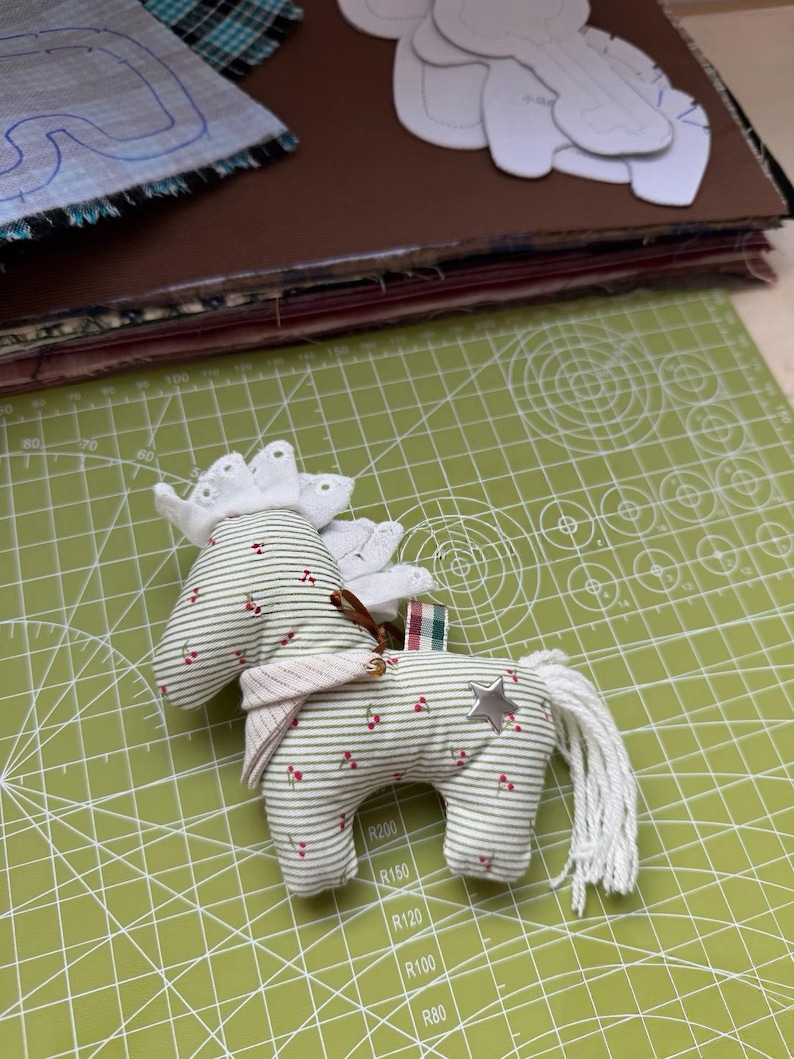 May include: A handmade fabric horse toy with a white mane and tail, adorned with a silver star and red berry accents. The horse is made of striped fabric with a white lace crown and a pink scarf. It rests on a green cutting mat.