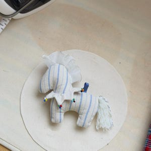 May include: A handmade toy horse made of striped fabric in white and light blue. The horse has a white mane and tail, a blue ribbon, and colorful bead accents. The toy is placed on a white circular surface.
