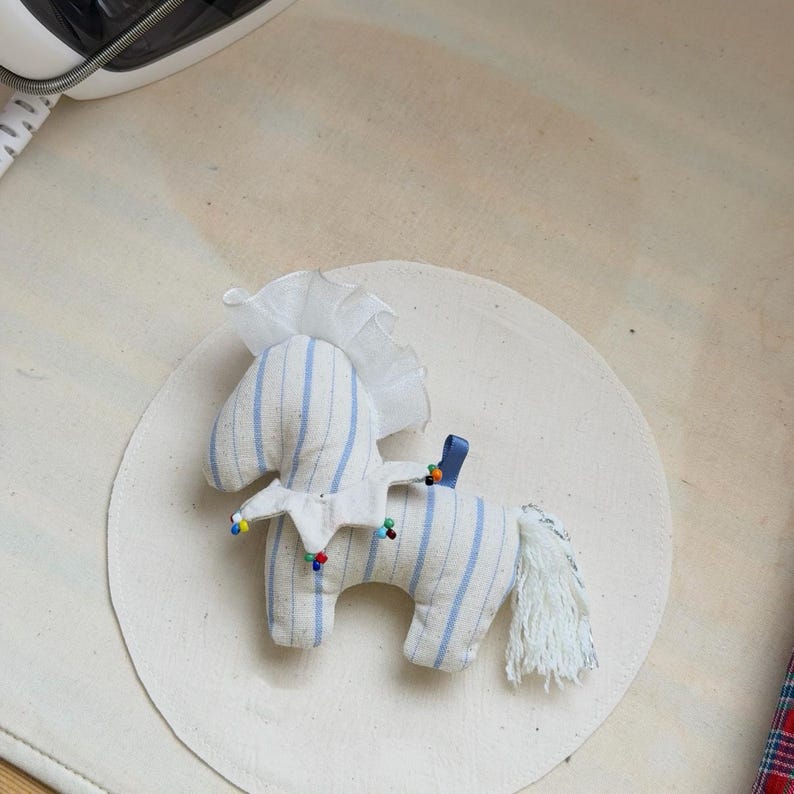May include: A handmade toy horse made of striped fabric in white and light blue. The horse has a white mane and tail, a blue ribbon, and colorful bead accents. The toy is placed on a white circular surface.