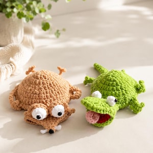 May include: Two crocheted animal figures: a tan creature with large white eyes and a small black nose, and a green alligator with white eyes, a red mouth, and a pink tongue. Both figures have textured yarn surfaces and small limbs.