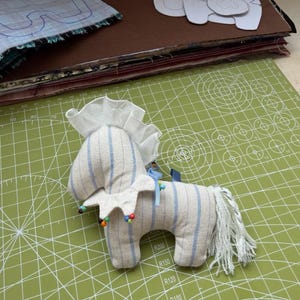 May include: A handmade stuffed horse toy with blue striped fabric and a white mane and collar. The toy is decorated with colorful beads and sits on a green cutting mat. Sewing patterns and fabric scraps are in the background.