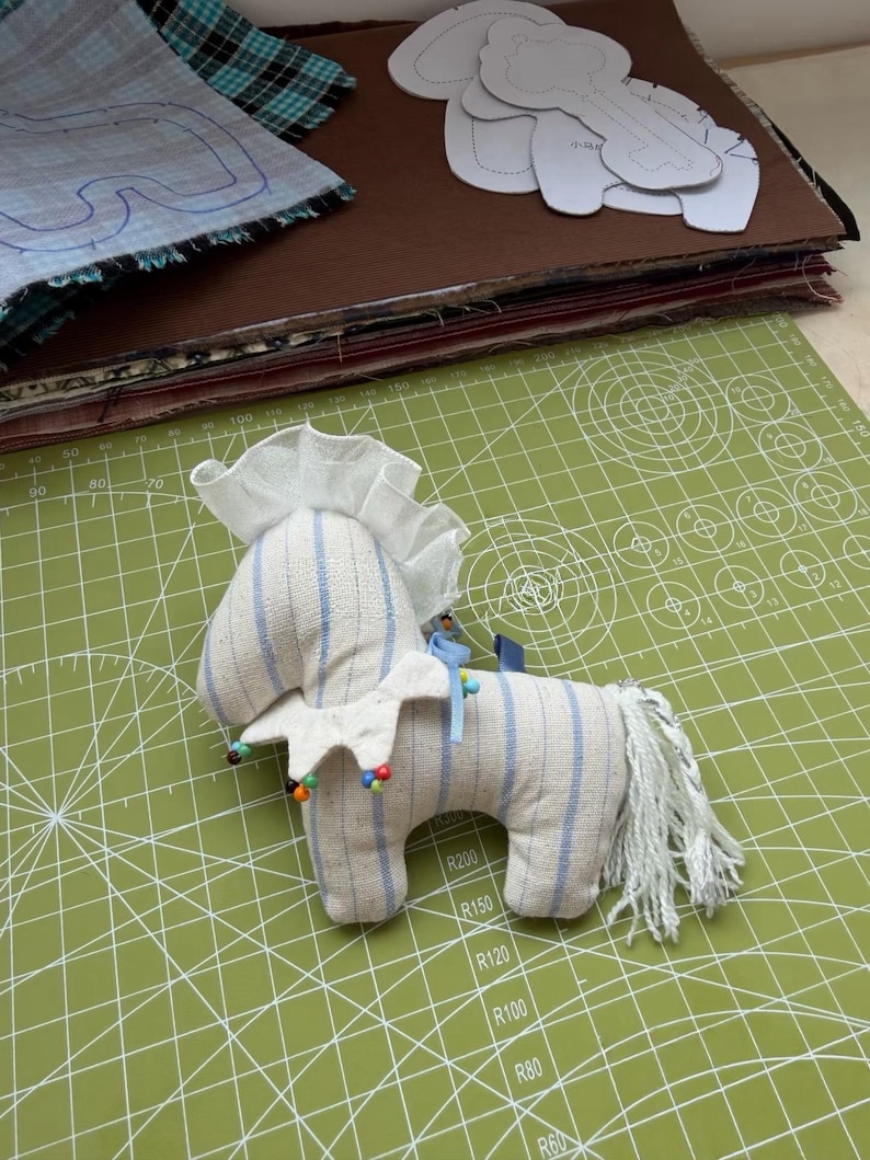 May include: A handmade stuffed horse toy with blue striped fabric and a white mane and collar. The toy is decorated with colorful beads and sits on a green cutting mat. Sewing patterns and fabric scraps are in the background.