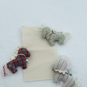 May include: Three handmade fabric horse ornaments in red plaid, green striped, and blue striped patterns. Each horse has a white mane and tail, and is decorated with small embellishments. The ornaments are displayed on a white surface.