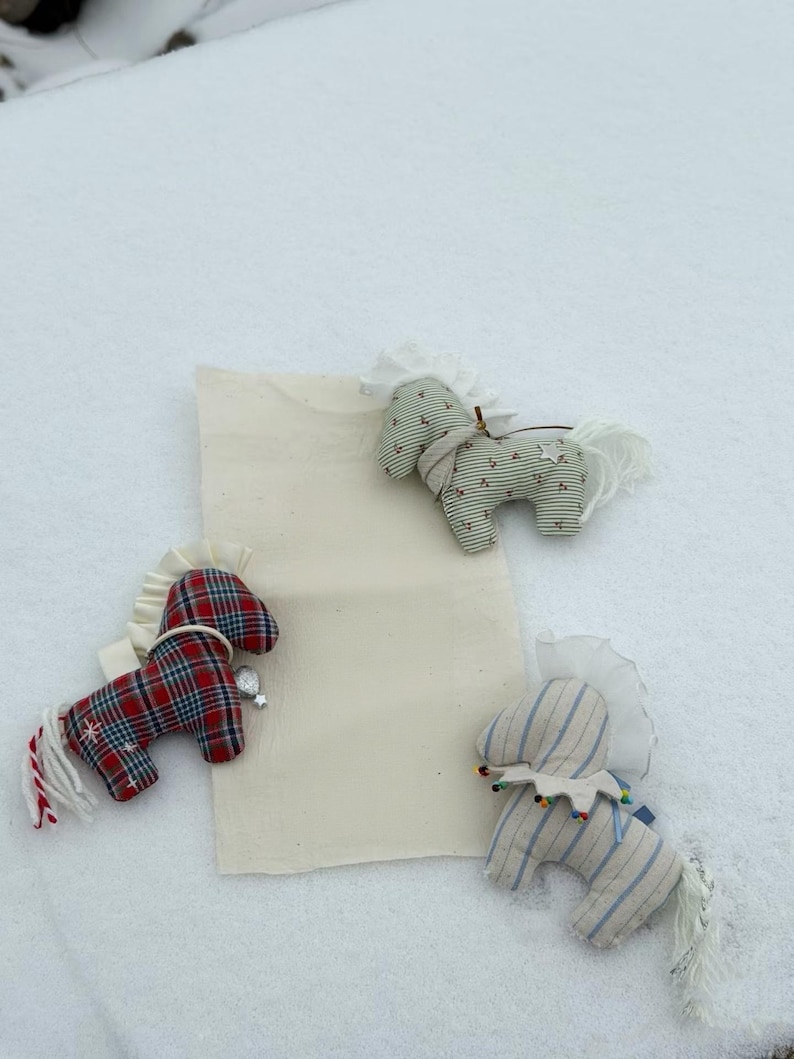 May include: Three handmade fabric horse ornaments in red plaid, green striped, and blue striped patterns. Each horse has a white mane and tail, and is decorated with small embellishments. The ornaments are displayed on a white surface.