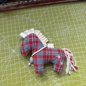 May include: A handmade fabric horse ornament with a red, blue, and white plaid pattern. It has a white mane and tail with red accents, a silver charm, and a white star detail. The ornament is on a green cutting mat.