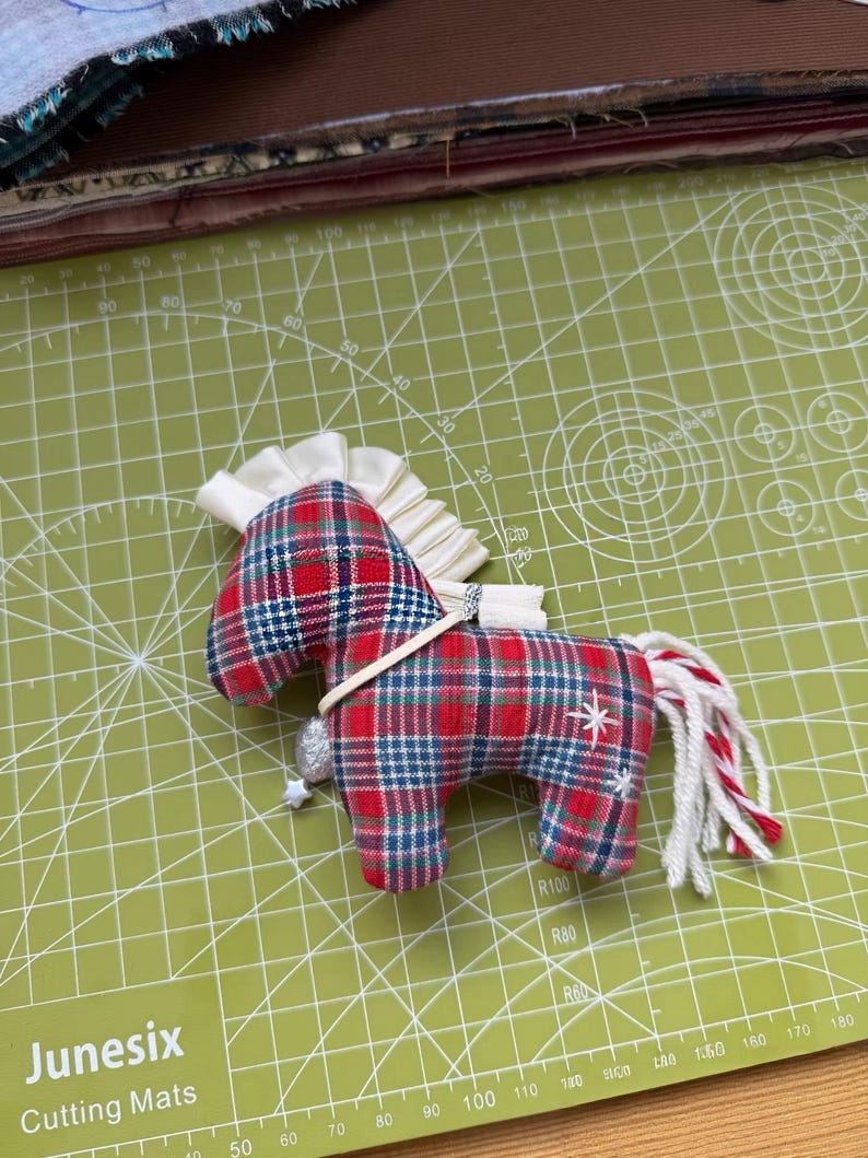 May include: A handmade fabric horse ornament with a red, blue, and white plaid pattern. It has a white mane and tail with red accents, a silver charm, and a white star detail. The ornament is on a green cutting mat.
