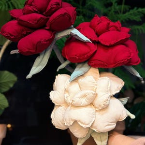 May include: A bouquet of three fabric flowers featuring two deep red blossoms and one cream-colored blossom. Each flower is constructed with multiple stuffed, rounded petals and includes light green fabric leaves attached to the stems.