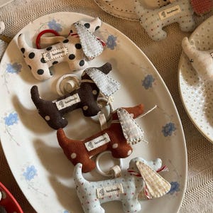 Puede incluir: Adornos de perro salchicha hechos a mano en varios patrones y colores, incluyendo lunares y estampados florales. Cada adorno tiene una pequeña etiqueta que dice "handmade" y está unido a un anillo.
