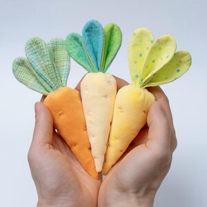 Op de afbeelding: Drie pluche wortels in tinten oranje, crème en geel, elk met groene stoffen toppen. De wortels worden in komvormige handen tegen een witte achtergrond gehouden. De stoffen toppen hebben verschillende patronen.