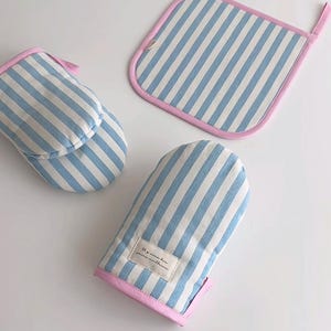 Może przedstawiać: Zestaw tekstyliów kuchennych z niebiesko-białym wzorem w paski. Zestaw zawiera rękawicę kuchenną, podkładkę pod garnek i maskę do spania, wszystkie obszyte różem. Rękawica kuchenna ma małą etykietę z tekstem.