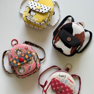 May include: Four small, colorful backpacks with various patterns and designs. One is yellow with a Snoopy design, another has a cowhide pattern, a third features a cat print, and the last one says "happy Camper".