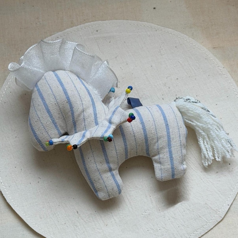 May include: A handmade stuffed horse toy. The horse is made of white fabric with blue stripes. It has a white mane and tail, and a decorative crown with colorful beads. The toy is on a white circular surface.