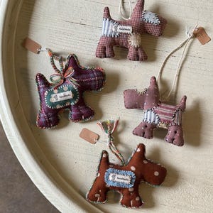 Puede incluir: Cuatro adornos de perro de tela hechos a mano en varios patrones y colores, incluyendo cuadros y lunares. Cada adorno tiene una pequeña etiqueta y una cuerda para colgar. Los adornos están dispuestos sobre una superficie redonda de color claro.