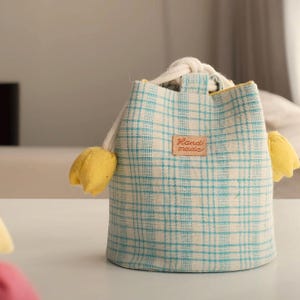 Pode incluir: Uma pequena bolsa feita à mão com um padrão xadrez azul e branco. A bolsa tem um fecho de cordão e é adornada com detalhes em forma de tulipa amarela. Uma pequena etiqueta retangular diz "Hand made".