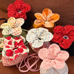 May include: A collection of drawstring pouches shaped like flowers with five rounded petals. The pouches feature various patterns including red with white dashes, orange plaid, multi-coloured polka dots, strawberry prints, and pink with white polka dots.