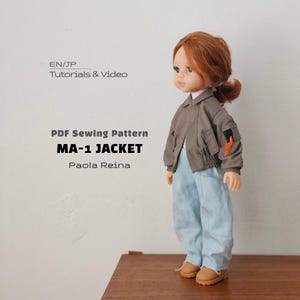 Könnte beinhalten: Eine Puppe mit rotbraunem Haar trägt eine graue MA-1-Jacke mit einer kleinen schwarzen Tasche und einem orangefarbenen Anhänger am Ärmel, dazu eine hellblaue Hose und hellbraune Schuhe. Text: EN/JP TUTORIALS & VIDEO PDF Sewing Pattern MA-1 JACKET Paola Reina.
