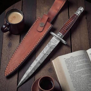 Puede incluir: Una daga de acero de Damasco con mango de madera marrón y funda de cuero. La daga está sobre una mesa de madera junto a una taza de café y un libro abierto. La hoja tiene un patrón detallado.