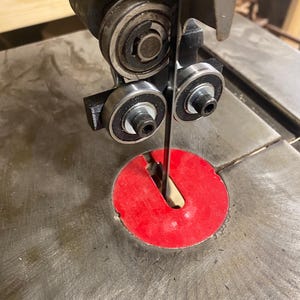 May include: Close-up of a band saw with a silver blade and black and silver components. The blade is positioned through a red insert on a metal table. The saw is designed for woodworking and cutting materials.