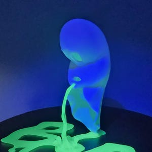 May include: A 3D printed figurine of a ghost-like figure glowing in blue and green under UV light. The figure is emitting a stream of bright green liquid that pools on the base. The text FTJ Astro is printed in the bottom right corner.