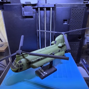 May include: A 3D printed olive green CH-47 Chinook helicopter model with black rotor blades, mounted on a black rectangular stand labelled CH-47 Chinook. The model features visible layer lines and is positioned in front of a 3D printer.