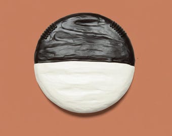 May include: A round, two-toned decorative object. The top half is black with a textured surface, and the bottom half is white. A black, ridged detail runs along the top edge. The object is set against a solid, muted orange-brown background.