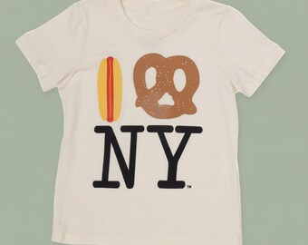 May include: Cream-colored t-shirt featuring a graphic design with a hot dog, pretzel, and the text "I NY" in black. The hot dog is yellow and red, and the pretzel is brown. The shirt is made of a soft material.