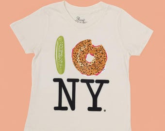 May include: Off-white t-shirt with a graphic design. The design includes a green pickle, a bagel with a bite taken out, and the text "I NY" in black. The shirt has short sleeves and a crew neck.