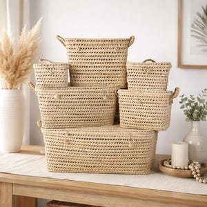 Puede incluir: Un conjunto de seis cestas de mimbre tejidas de varios tamaños, apiladas sobre una mesa de madera. Las cestas son de color beige natural con asas y un patrón texturizado. Adecuado para almacenamiento o decoración.
