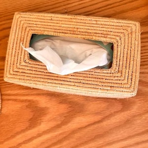 Könnte beinhalten: Ein rechteckiger Taschentuchbox-Bezug aus geflochtenen Naturfasern. Der Bezug hat eine rechteckige Öffnung und ist mit weißen Taschentüchern gefüllt. Die Box steht auf einer Holzoberfläche.