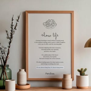 May include: A framed print with the words "slow life" in a minimalist style. The print is in a light wood frame and is displayed with decorative vases, a small cactus, and a brass pendant light.