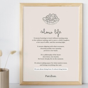 May include: A framed print with the words "slow life" in a handwritten style. The print has a beige background with black text and a line drawing of a flower. A vase with dried flowers is in the foreground.