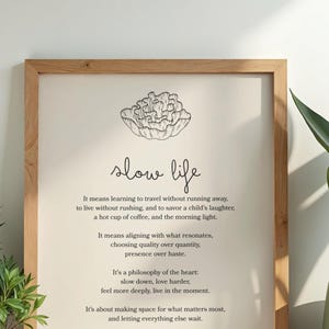 May include: Wooden framed print with the words "slow life" in a handwritten font. The print includes a floral illustration and text about slowing down and appreciating life's simple pleasures. A potted plant is visible on the right.