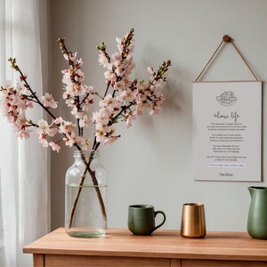 May include: A wooden table displays a clear glass jar with cherry blossom branches, a green mug, a gold-colored cup, and a green pitcher. A framed print with the words "slow life" hangs on the wall.