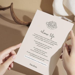 May include: A beige card with the words "slow life" in script, a floral illustration, and text about slowing down. The card is held by two hands, with a brown antler and fabric in the background.