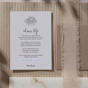 May include: A light gray card with the words "slow life" and a floral illustration. The card has French text and is set against a textured background with neutral tones, including beige and brown.