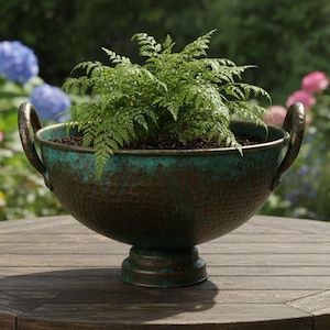 Puede incluir: Una maceta de bronce con helechos verdes. La maceta tiene dos asas curvas y una base de pedestal. Se encuentra sobre una mesa redonda de madera en un jardín con flores.