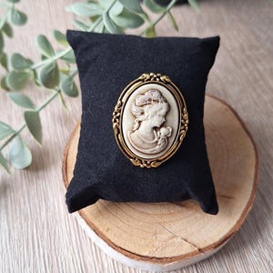 Peut inclure: Broche camée ovale avec un cadre doré, représentant le profil d'une femme. Le camée est posé sur un coussin en velours noir, lui-même posé sur une tranche de bois. Le fond comprend de la verdure.