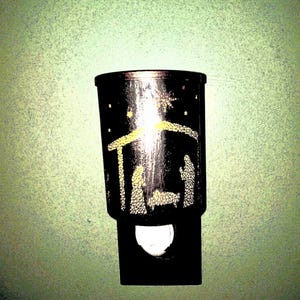 May include: Black night light with a nativity scene design. The cylindrical night light features a silhouette of the holy family and a star. The base of the night light has a white, semi-circular light.