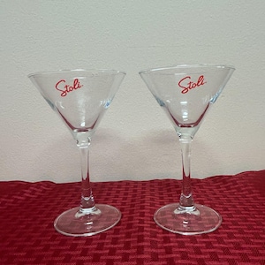 以下が含まれることがあります： 赤い文字で「Stoli」と書かれた2つの透明なマティーニグラス。グラスは三角形のボウル型で、長いステムが付いています。赤い模様の表面に置かれています。