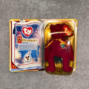 Puede incluir: Un peluche Beanie Baby Millennium the Bear en su embalaje original. El oso burdeos tiene una cinta dorada y un parche 2000. El embalaje incluye un folleto con el logotipo de McDonald's y el texto "Ronald McDonald House Charities".